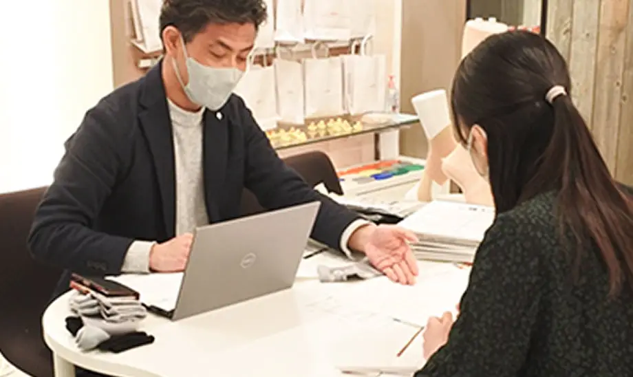 マスクをつけた人物がデスクでノートパソコンを使用しながら相談している様子。