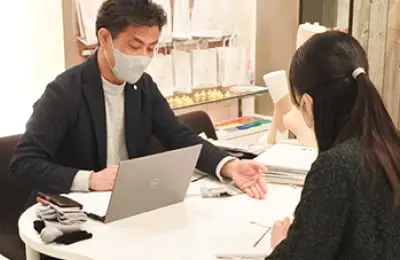 マスクをつけた人物がデスクでノートパソコンを使用しながら相談している様子。