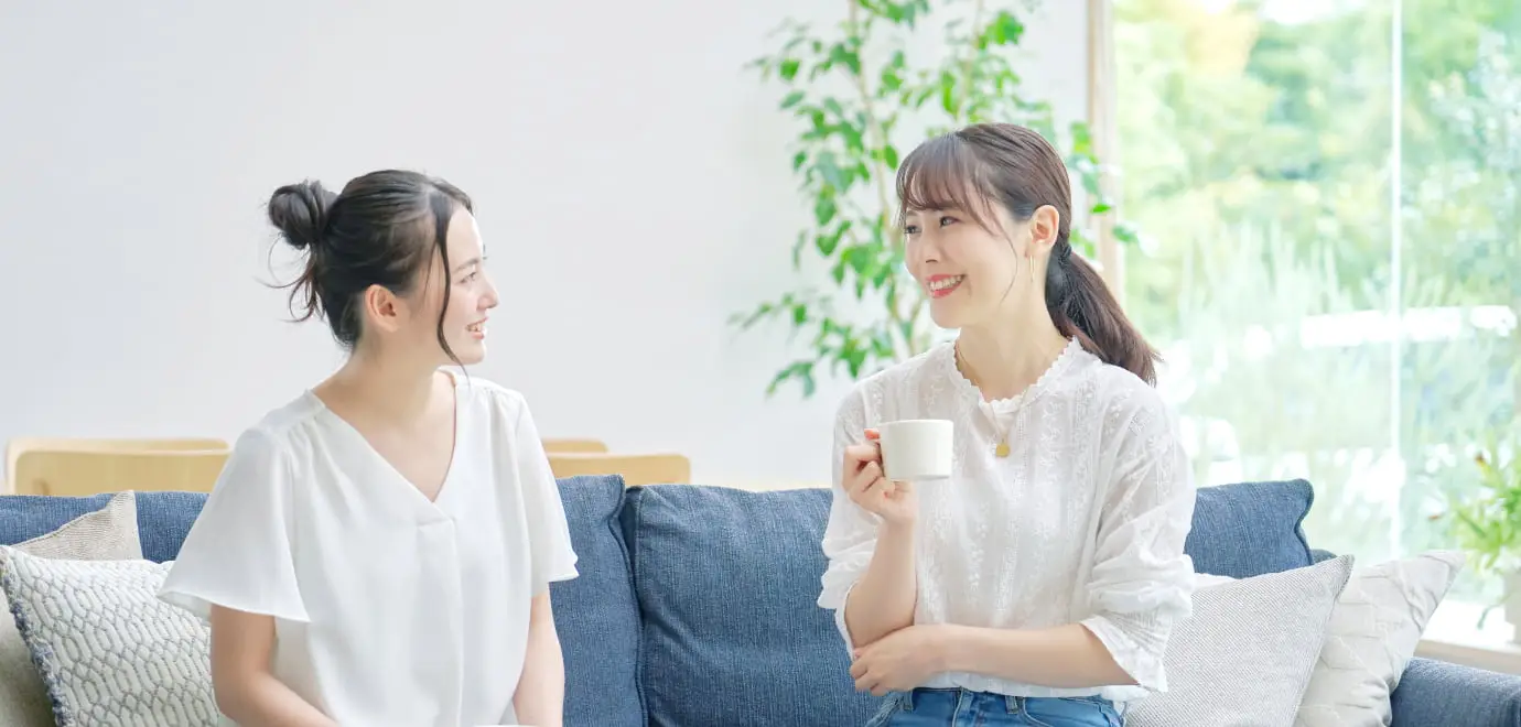 明るいリビングルームで、白い服を着た二人の女性がソファに座って会話している様子。