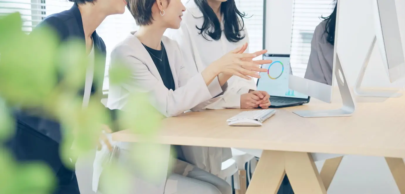 オフィスで白いジャケットを着た人物がパソコンの前で説明している様子。