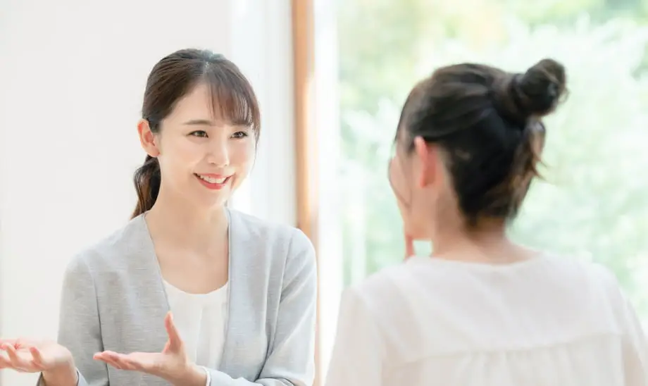 明るい室内で、グレーのジャケットを着た女性が笑顔で相手と会話している様子。